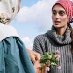 Two women outdoors in headscarves holding wildflowers under a clear sky.
